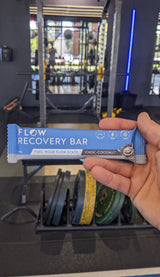 Hand holding a Flow Recovery Bar in front of gym equipment